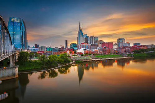 City skyline at sunset with reflective river and bridge in foreground.