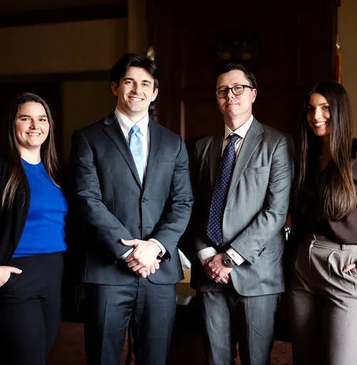 Four professionally dressed individuals standing indoors, smiling, two men in suits and two women in business attire.