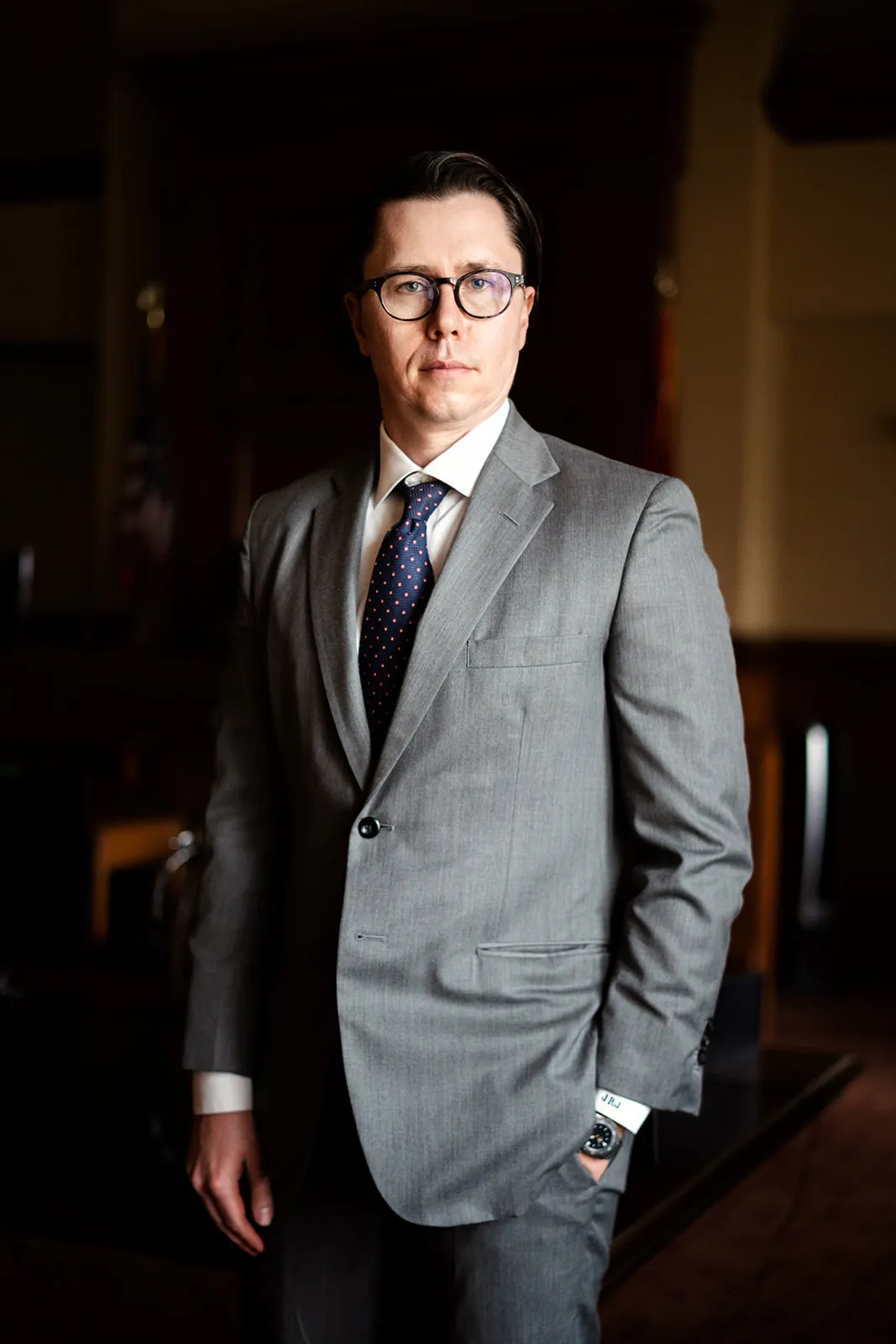Professional man wearing a gray suit, white shirt, navy tie, and glasses standing indoors.