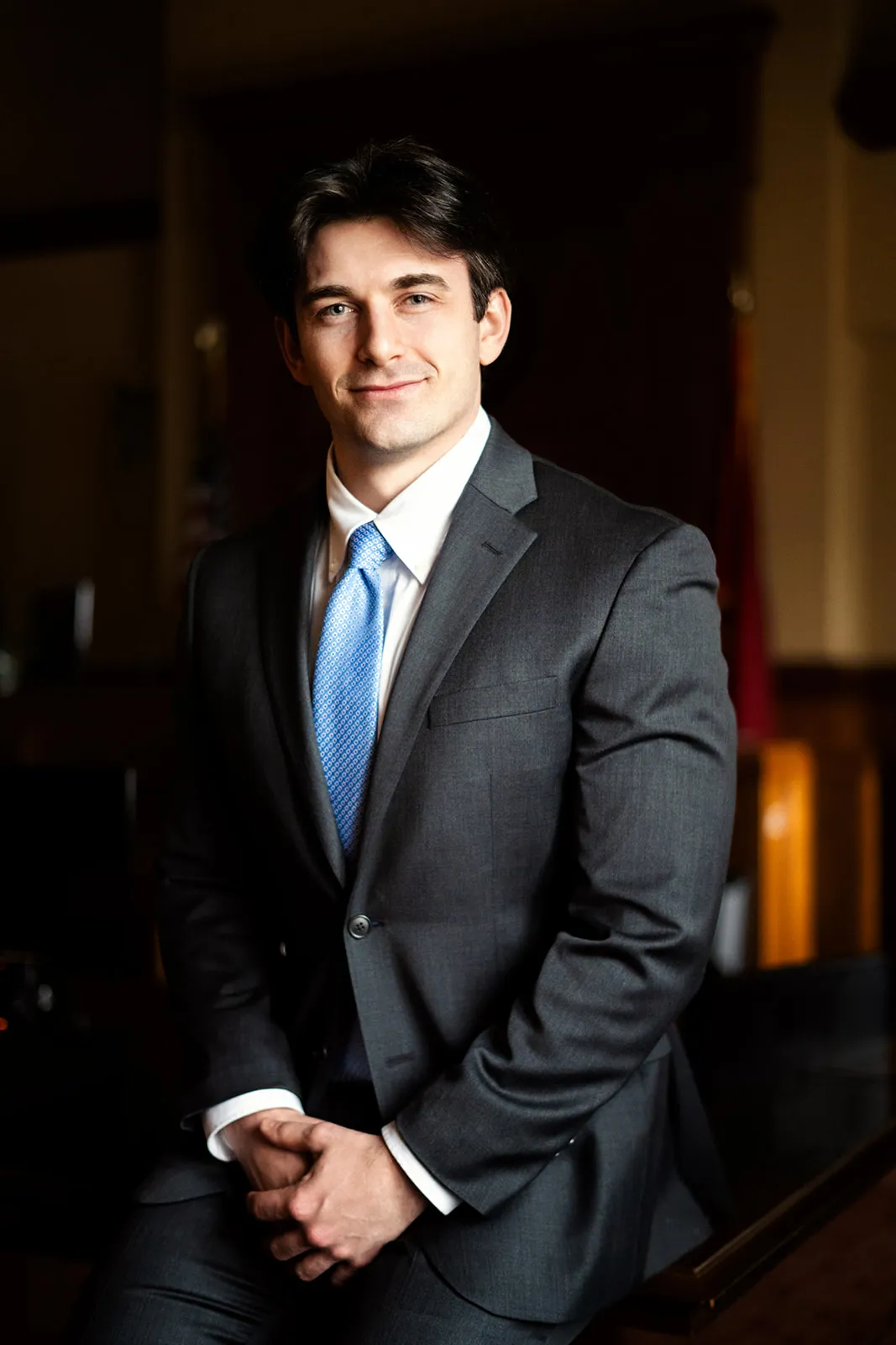 Portrait of a young man in a dark gray suit and blue tie, seated indoors.