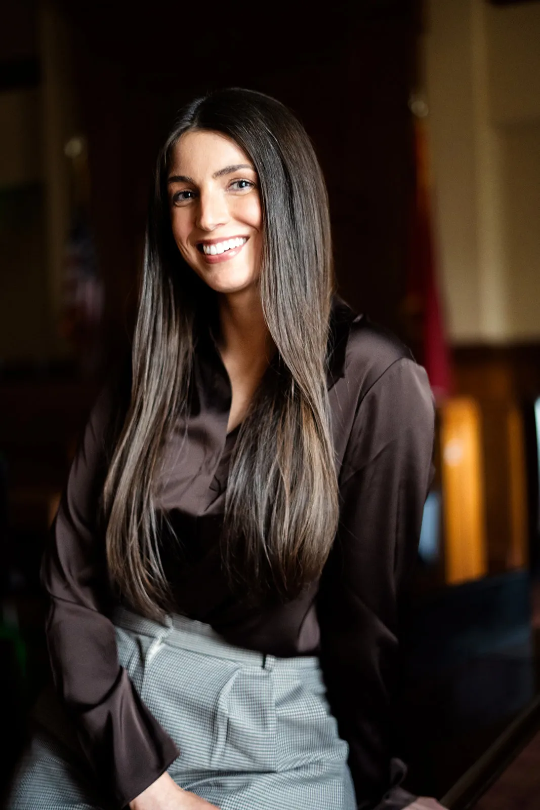 Portrait of a smiling woman with long dark hair wearing a brown blouse and gray pants.