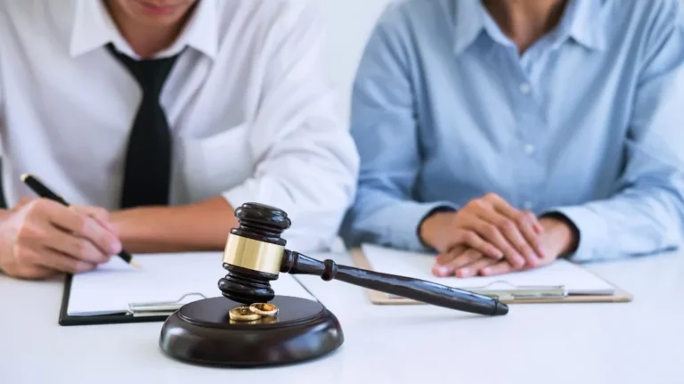 Two individuals in formal attire seated at a table with a judge's gavel and two gold wedding rings.