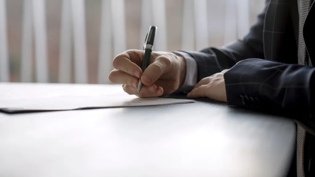 Close-up of a person in a suit signing a document with a black pen on a white surface.
