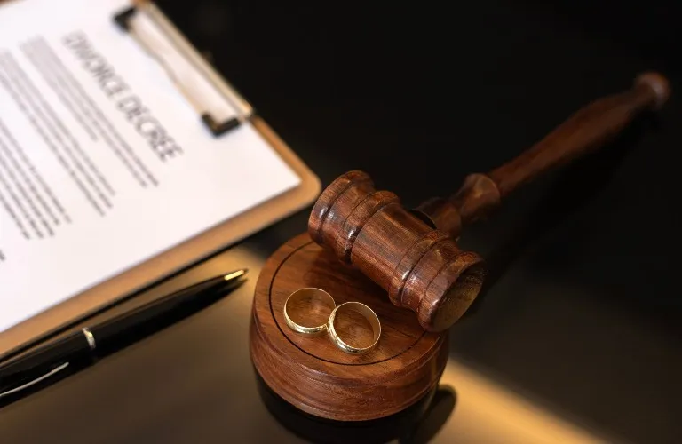 Wooden judge's gavel and base with two gold wedding rings, pen, and divorce decree document.