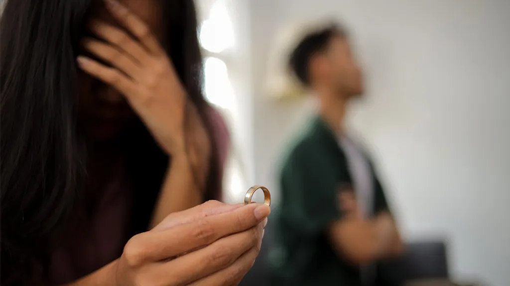 Woman holding a wedding ring with a distressed expression, man sitting blurred in background.