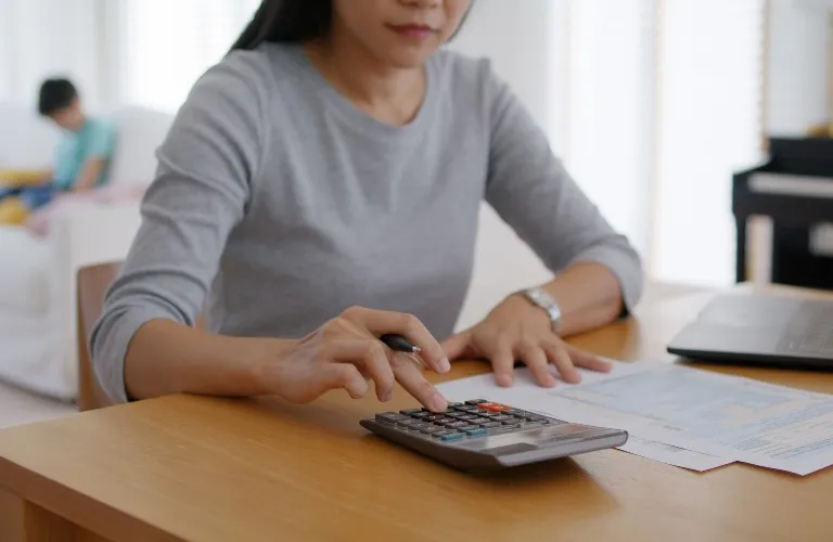 Woman in gray shirt using calculator at wooden table with documents and laptop.