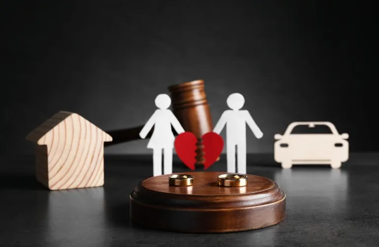 Wooden house and car models, paper cutout man and woman holding broken heart, judge's gavel, and two wedding rings on wooden platform.
