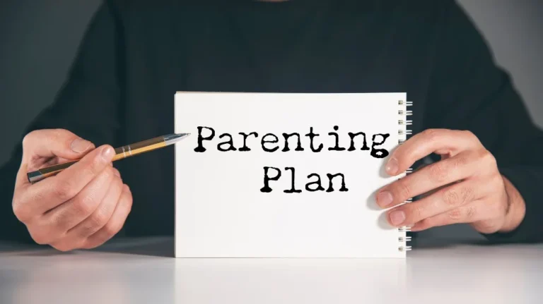 Person holding a white spiral notebook with "Parenting Plan" text and a pen.