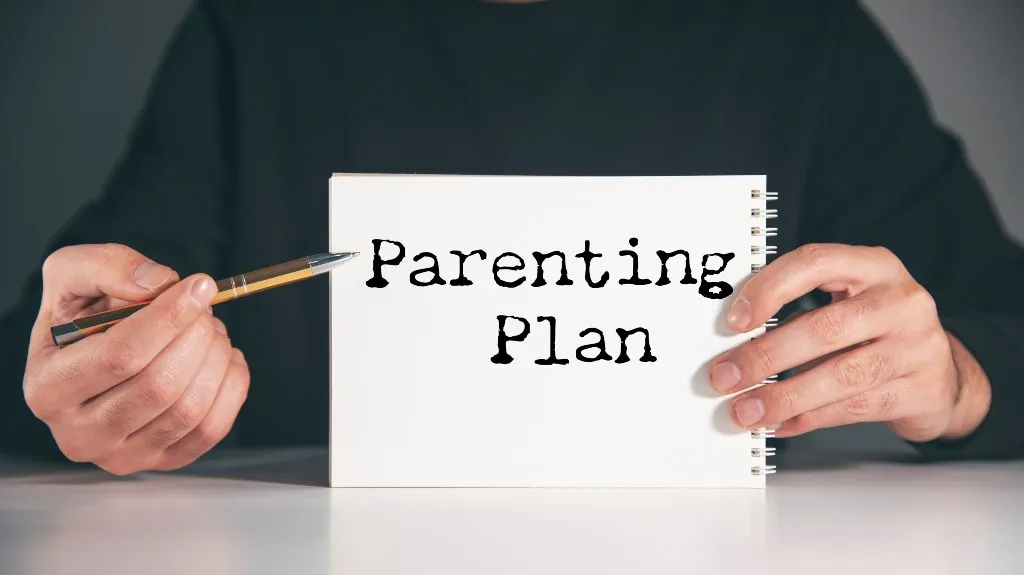 Person holding a white spiral notebook with "Parenting Plan" text and a pen.