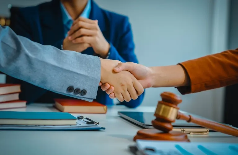 Two individuals in formal attire shaking hands across a desk with legal books and a judge's gavel.
