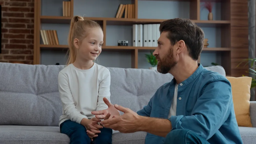 Man in blue shirt talking to smiling young girl on gray couch in living room.