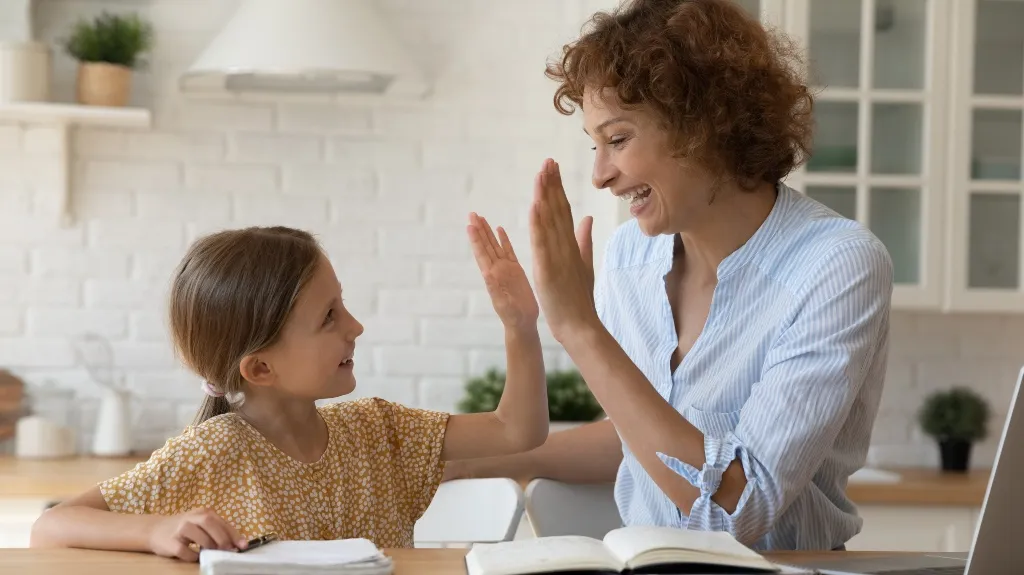 Woman and young girl giving a high-five in a bright kitchen with books on the table.