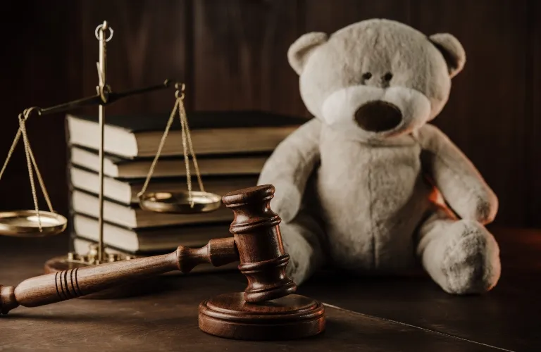 Wooden judge's gavel, brass scales of justice, stacked books, and a plush teddy bear on a dark surface.
