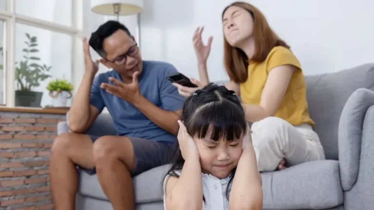 Young girl covering ears with hands while parents argue on a gray sofa indoors.