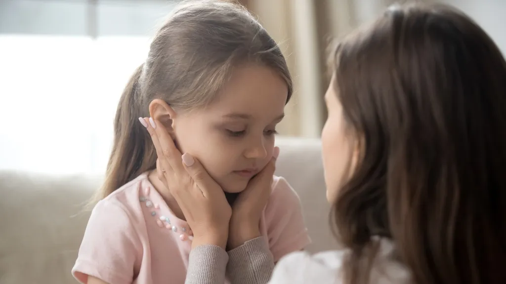 Adult woman gently holding and comforting a young girl with light brown hair indoors.